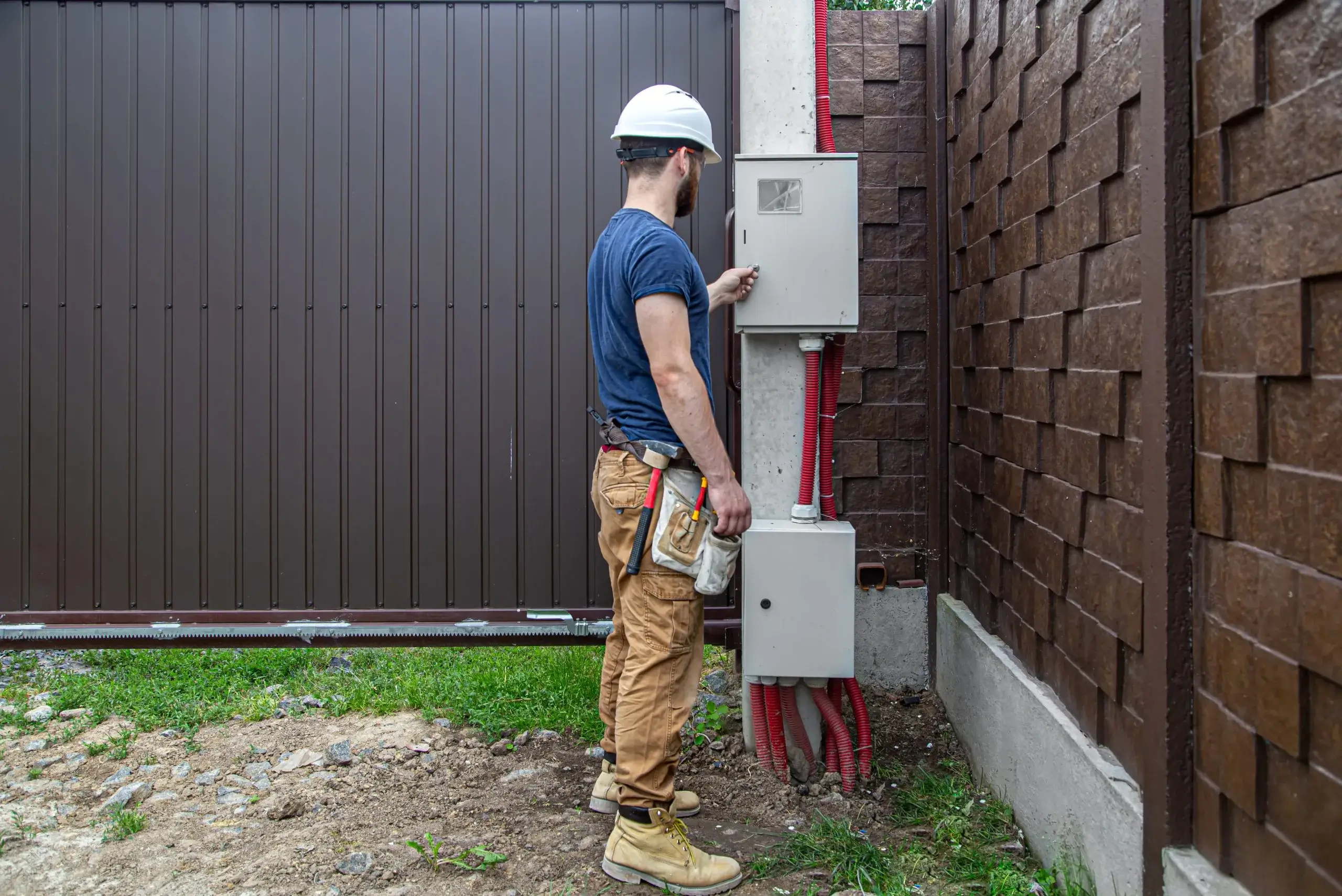 A construction worker in a hard hat inspects an electrical panel by a fence