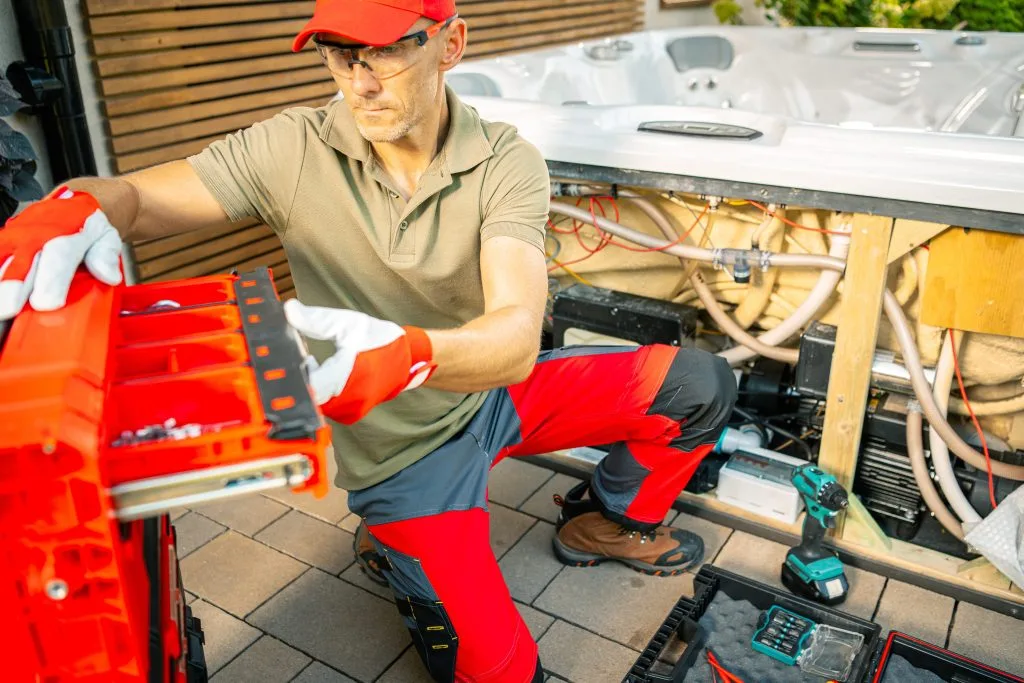 A technician in red pants and gloves repairs equipment beside a hot tub